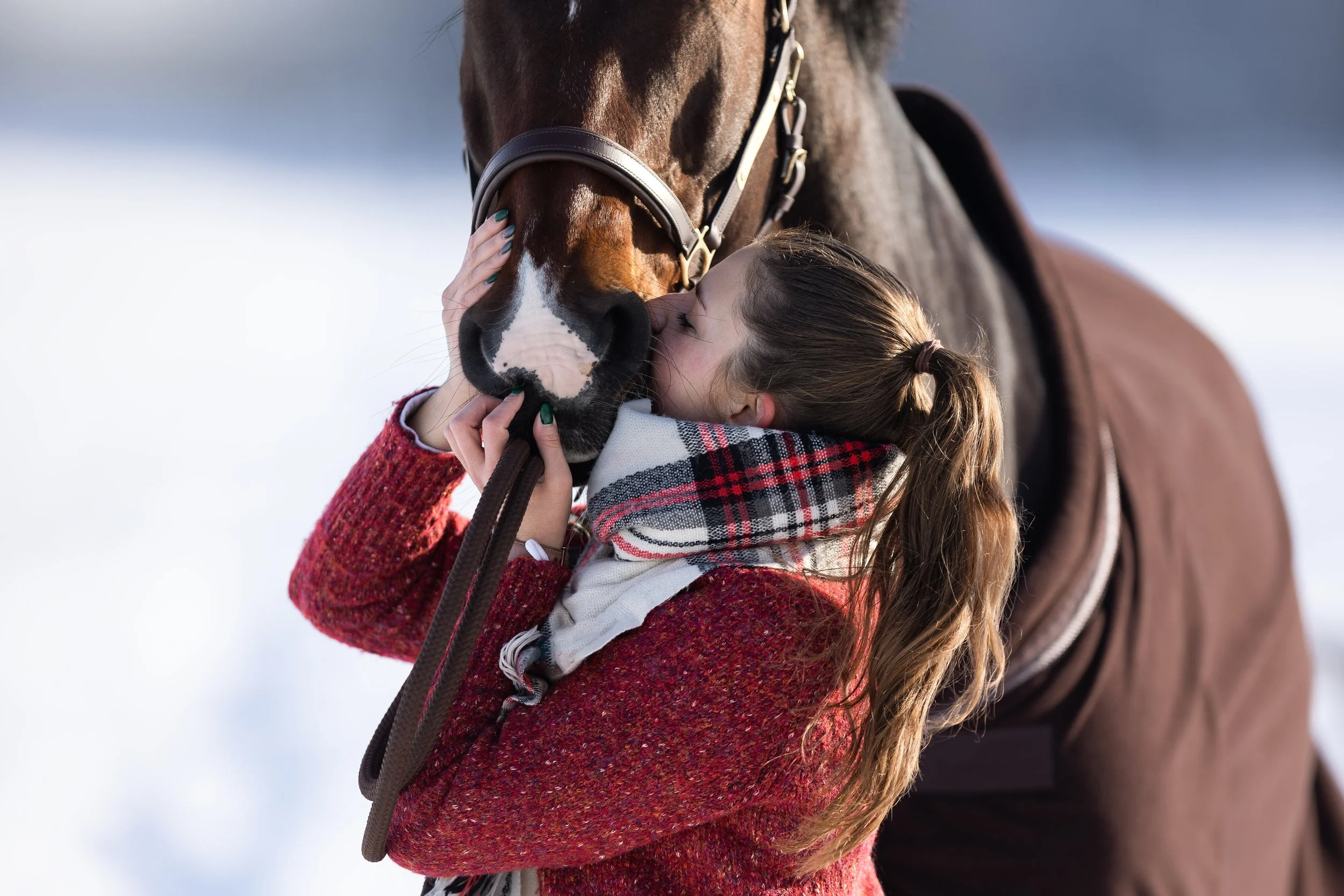 Žena objímá a líbá koně na čumák venku, se sněhem v pozadí.
