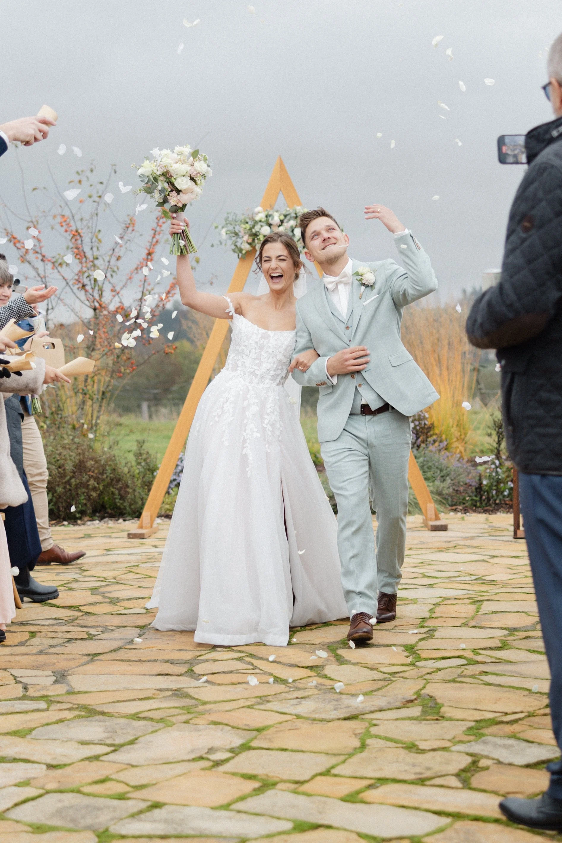 Novomanželský pár prochází uličkou venku, usmívá se a slaví, zatímco hosté házejí květinové lístky a konfety.