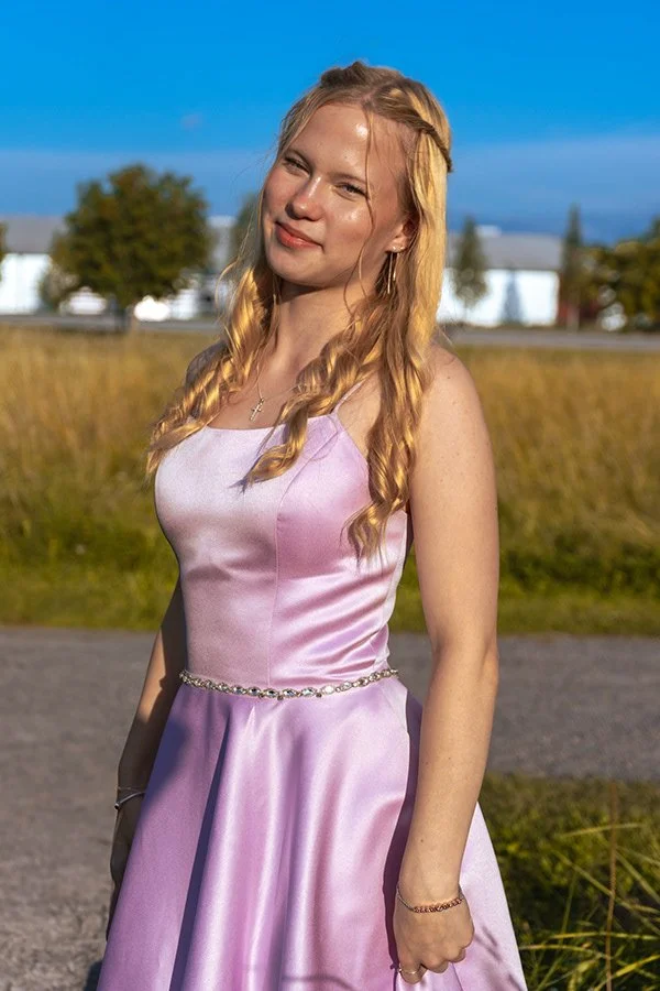 Nuori nainen hymyilee kevättakaisessa pikkutakissa, taustalla avoin maaseutu ja sininen taivas.