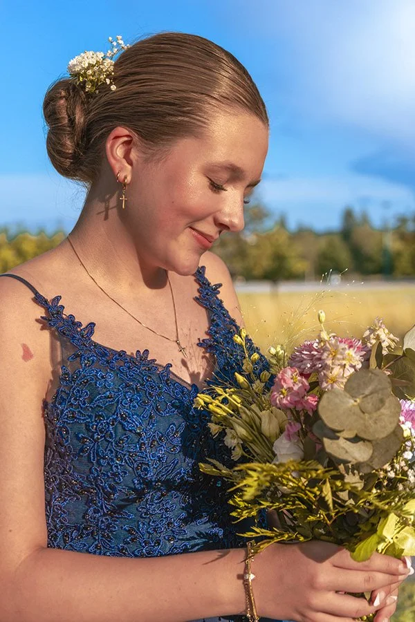 Nuori nainen on juhlapuvussa, pitää kukkakimppua, ja hymyilee varovasti. Taustalla on kirkas taivas ja avoin maisema.