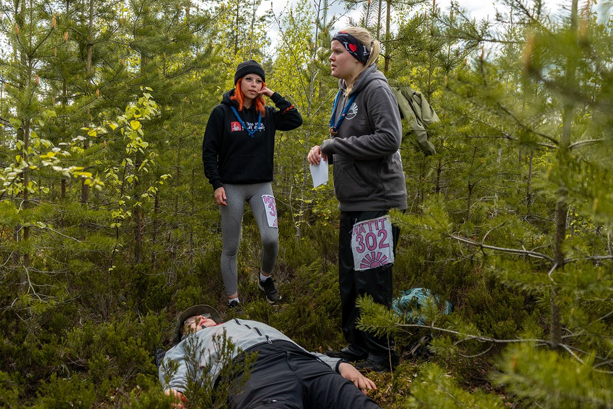 Kolme nuorta ihmistä metsässä, yksi makaa maassa tajuttomana, kaksi muuta keskustelee hänen kanssaan.