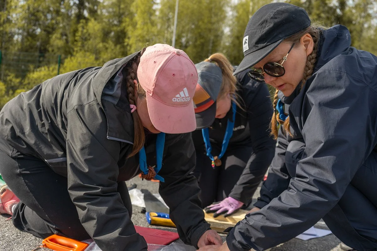 Kolme ihmistä polvistuu ja tekee ensiaputestiä ulkona, taustalla puita ja sininen taivas.
