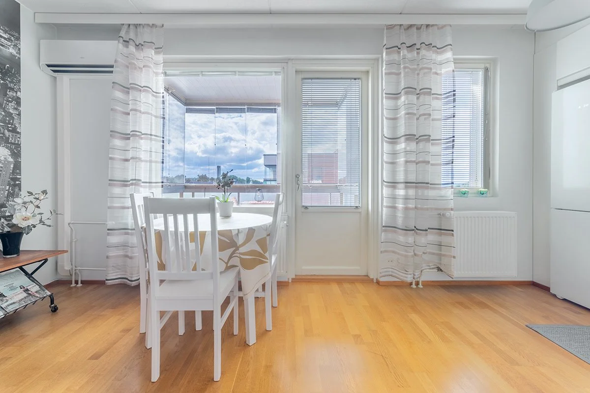 Valkea sisätila ruokapöydällä ja valkoiset tuolit, ikkunoissa ja ovessa valkoiset verhot, puulattia, lattialla pieni matto.