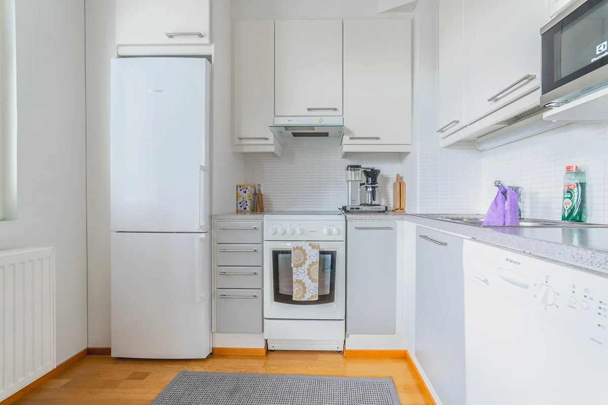 Kodin keittiö, valkoinen ja harmaa, jääkaappi, uuni, astianpesukone, mikroaaltouuni, jääpiippu, kahvinkeitin, pesupähkinäpuoli, pyyhe, tiskiharja, ja astiahylly.