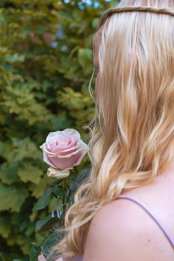 Nainen nähdään sivulta, hänellä on pitkät, kiharat vaaleat hiukset ja hän pitää ruusua, jonka hän on tuonut kasvin varresta, taustalla vihreitä lehtiä.