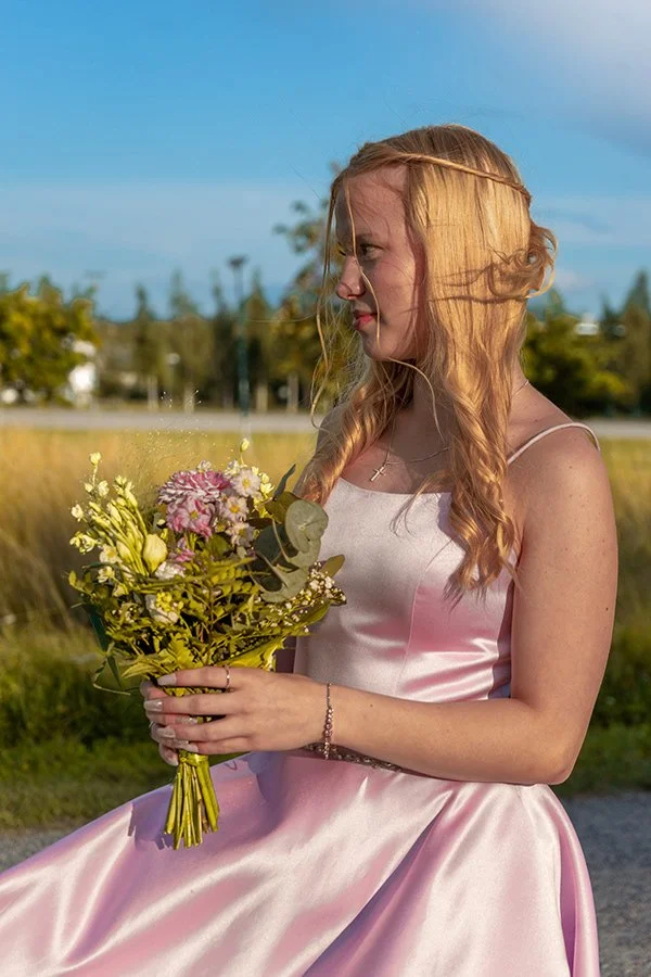 Nuori nainen istuu ulkona, pukeutuneena vaaleanpunaiseen satiinipukuun ja pitää kukkaseppeleen. Hänellä on pitkä, muodoltaan laineikas hius, ja taustalla on pelto ja puut sinisellä taivaalla.