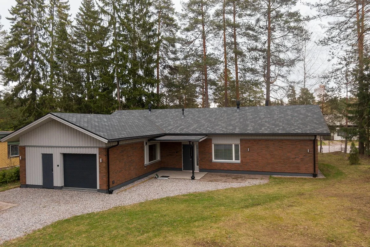 Yksi-tasoinen talo, jossa on autotalli, punatiilimuuri ja valkoiset ikkunat, sijaitsee pihapiirissä nurmikon ja hiekkakäytävän ympäröimänä, taustalla puita ja osittain pilvinen taivas.