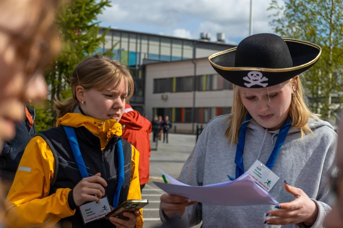 Nuorta ihmisiä ulkona. Toinen nainen pukeutunut piraattipäähineeseen katsoo papereita, toinen katsoo puhelinta.