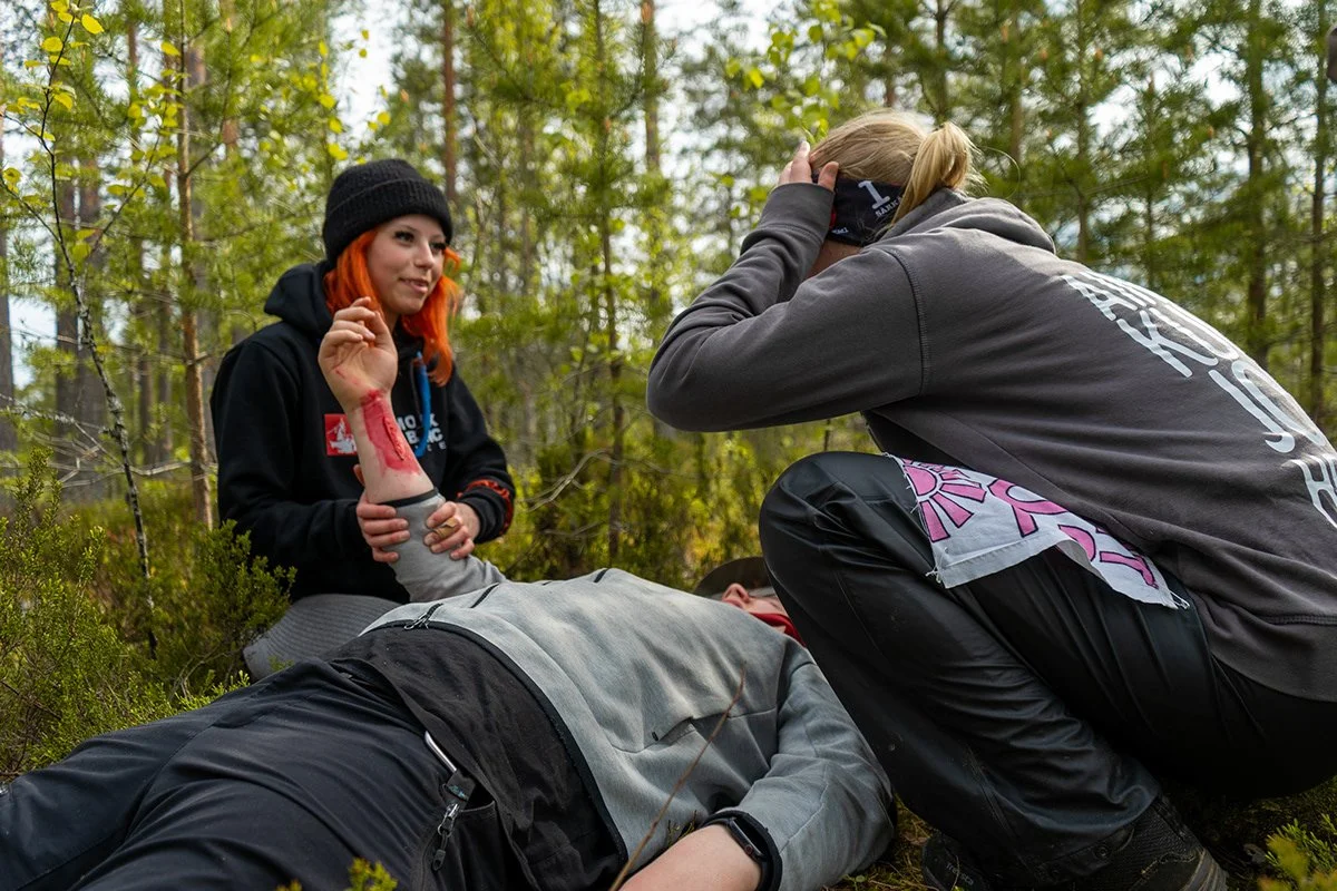 Kolme nuorta aikuista, kaksi naista ja yksi mies, auttavat loukkaantunutta henkilöä metsässä, yksi nainen antaa ensiapua ja toinen pitää tämän päätä, loukkaantunut makaa maassa.