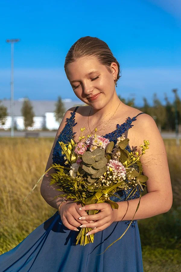 Nuori nainen hymyilee ja pitää kukkakimppua, hän on mekkossa ja iloisessa mielessä ulkona auringonpaisteessa.