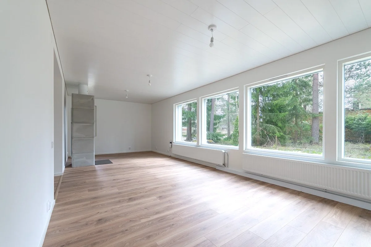 Tyhjä, valoisa olohuone suuret ikkunat ja puulattia.