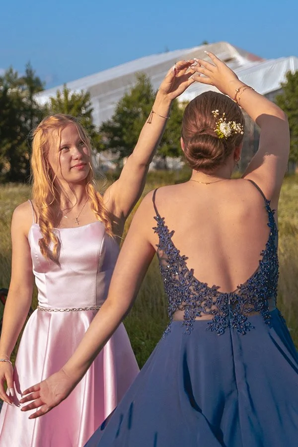 Kaksi nuorta naista tanssivat ulkona, toinen pukeutunut vaaleanpunaiseen ja toinen tummansiniseen pukuun, heidän kädet ovat toistensa yllä, taustalla vihreää luontoa ja sininen taivas.