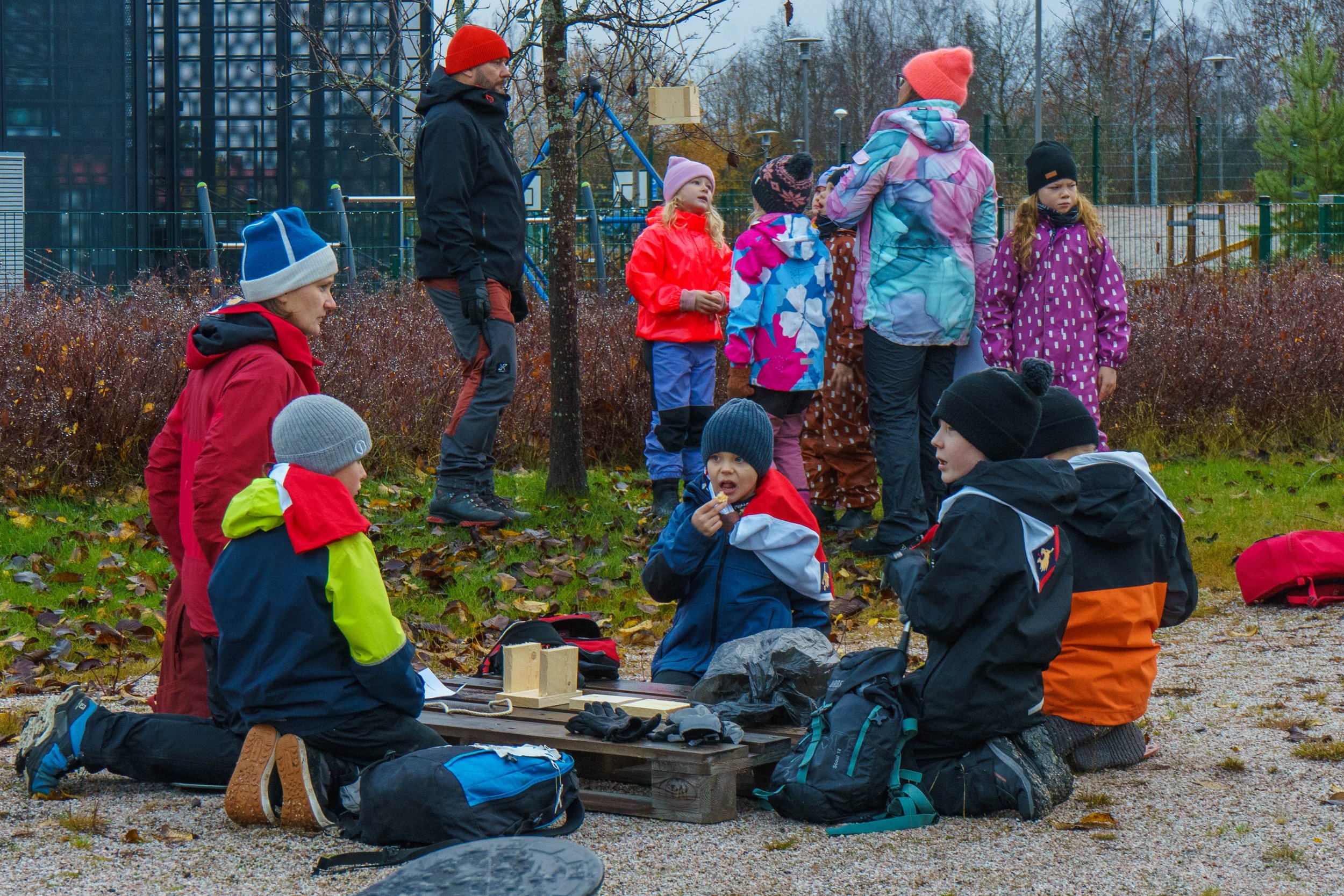 Ryhmäläisiä ulkona kerhotapahtumassa, lapset ja aikuiset istuvat ja seisovat nurmikolla, varustautuneet kylmään säähän, ympärillä puita ja pelialue, tummat pilvet taivaalla.
