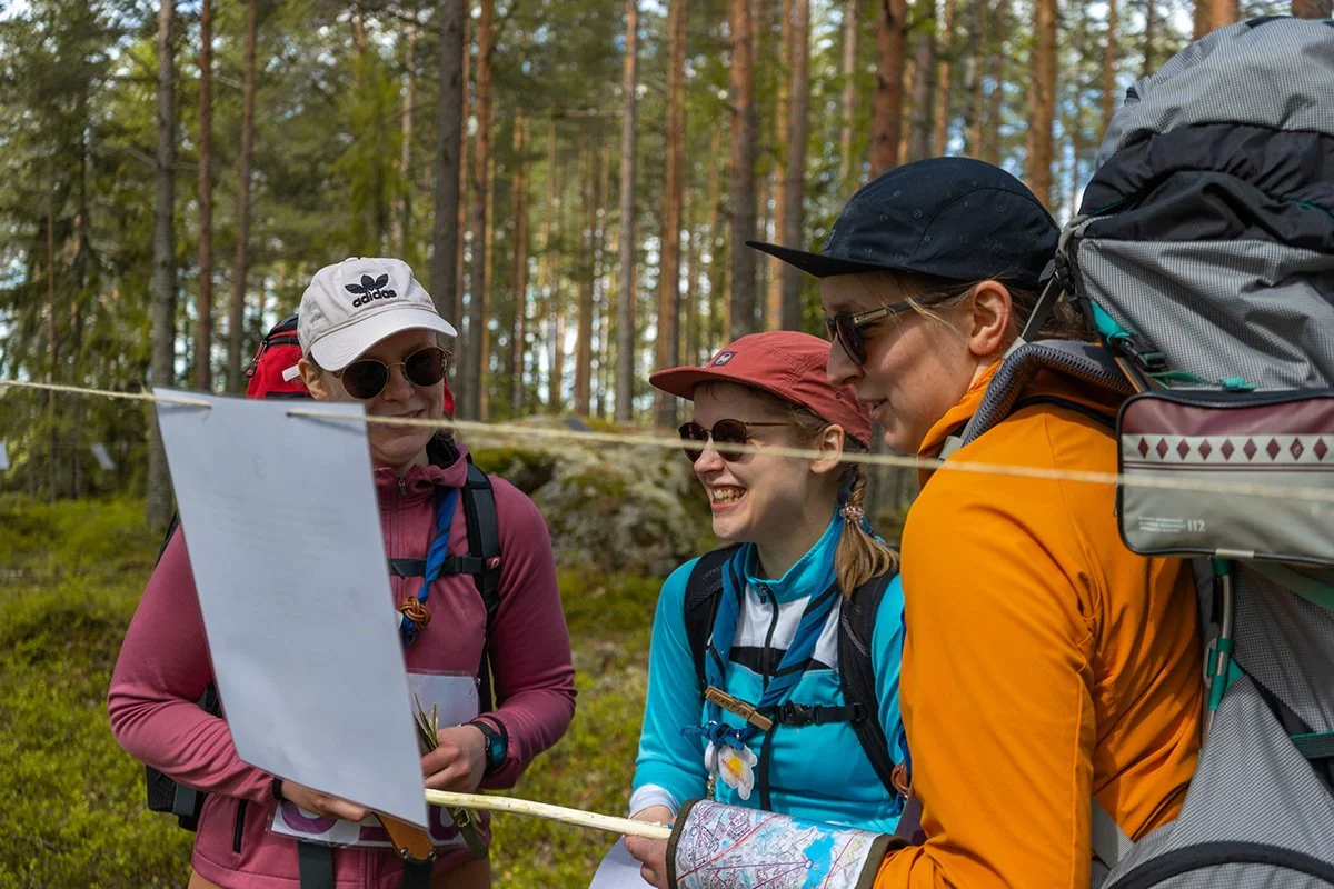 Kolme naista ulkona metsässä, juttelevat ja katsovat paperia tai karttaa, heillä on reppuja ja ulkoiluvarusteita