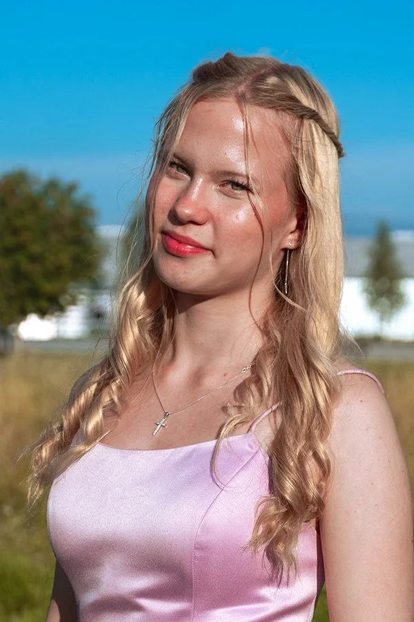 Nuoren naisen muotokuvalähikuva ulkona kirkkaassa auringonvalossa, taustalla puut ja yksi rakennus, hänellä pitkät vaaleat hiukset ja vaaleanpunainen mekko.