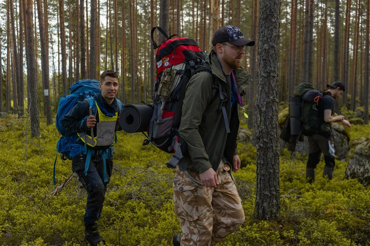 Kolm retkeilijää metsikössä, kaksi etualassa ja yksi taustalla, kantavat suuria reppuja ja varusteita.