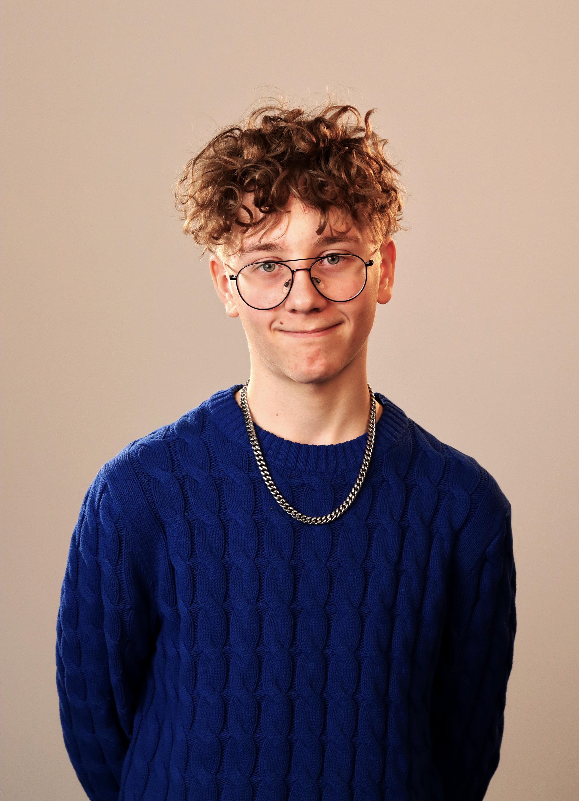 Nuori mies, silmälasit, khakki kihara hiukset, sininen villapaita, hopeinen ketju, neutraali tausta.