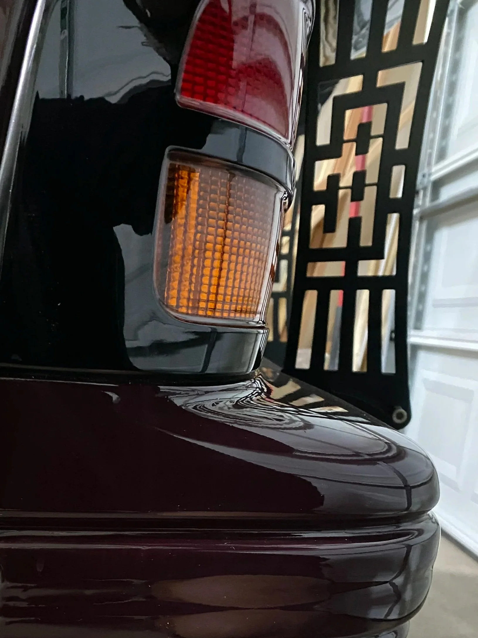 Close-up of the rear corner of a vintage black car, showing the tail light and part of the bumper, with a decorative black metal chassis mount wing and a garage door in the background.
