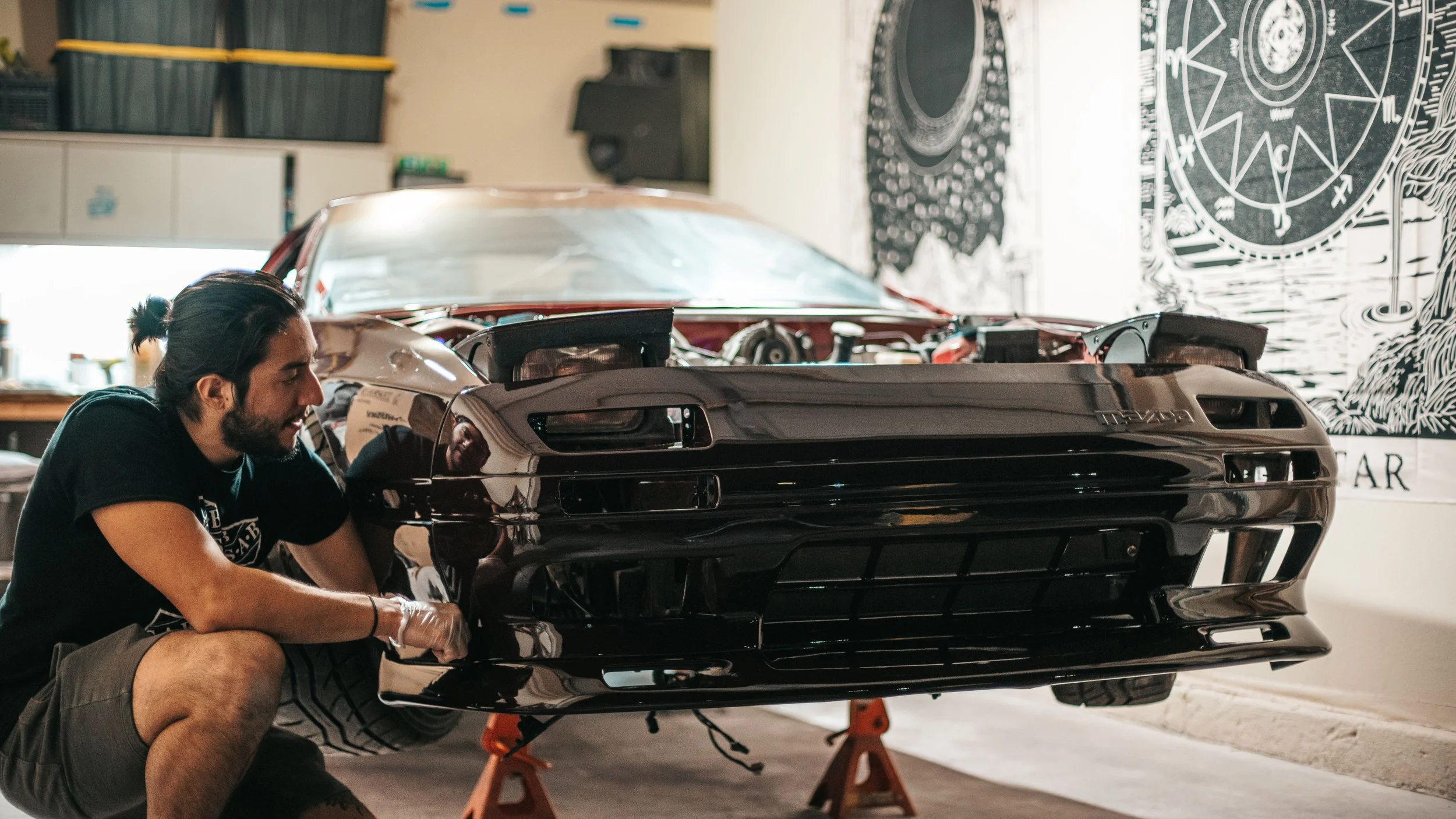 A man working on a black sports car inside a garage or workshop, with tools and storage bins in the background.