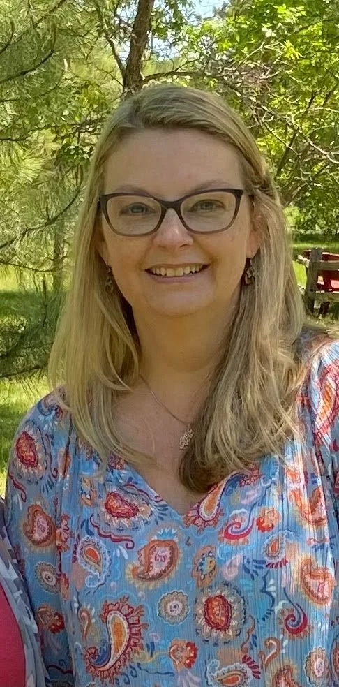 A woman with blonde hair wearing glasses, a patterned blue, red, and orange blouse, and earrings, smiling outdoors with trees and a pond in the background.