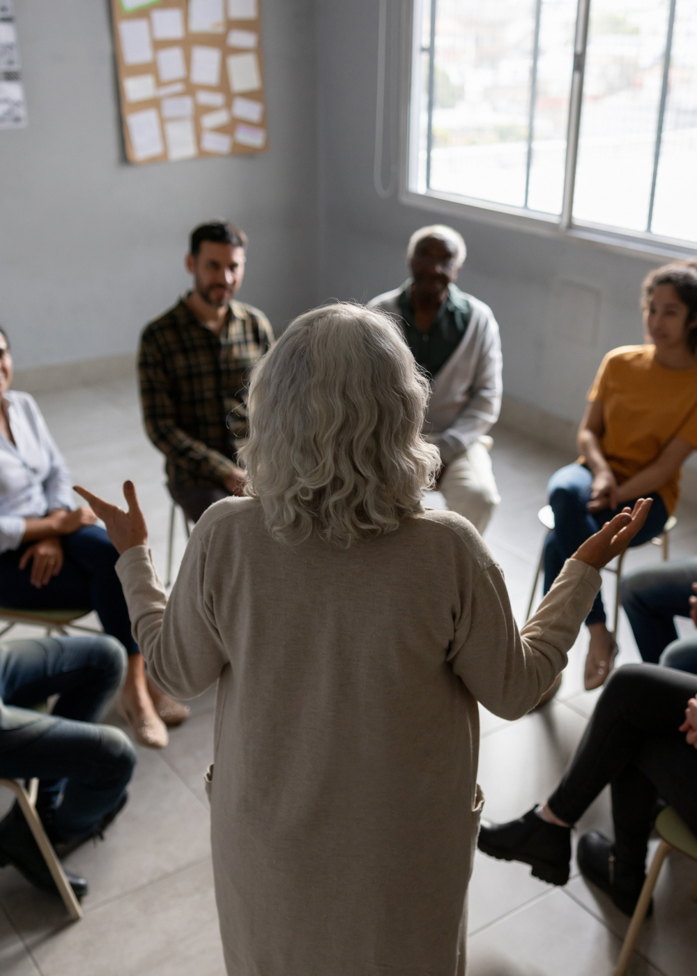 A woman with gray hair leading a group discussion in a bright room with large windows, a bulletin board, and several people seated in a circle around her.