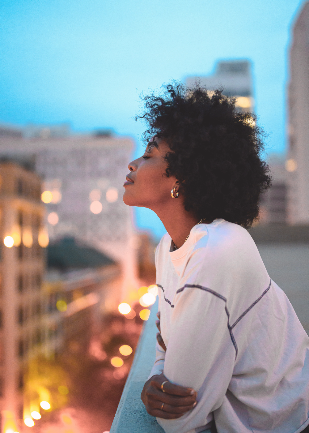 A woman with dark skin and curly black hair stands on a city balcony at dusk, eyes closed, looking peaceful with city lights and buildings blurred in the background.