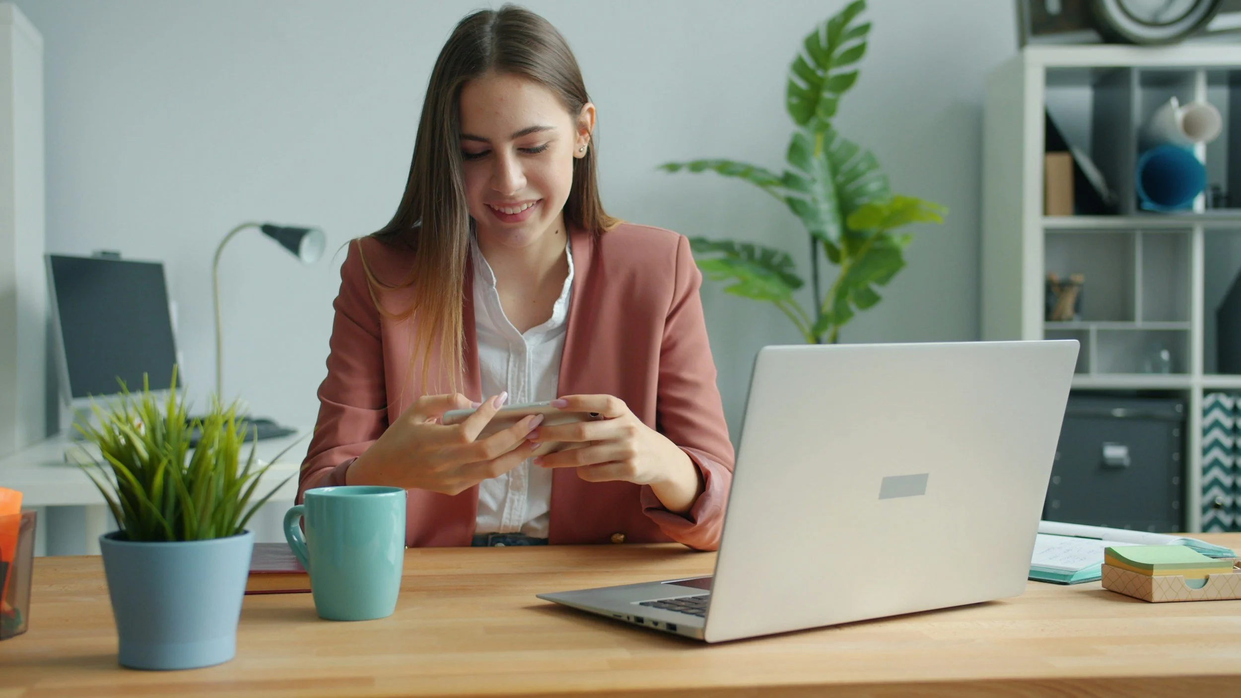 A woman sitting at a desk in an office, smiling while using her smartphone. The desk has a laptop, a potted plant, a coffee mug, and office supplies.