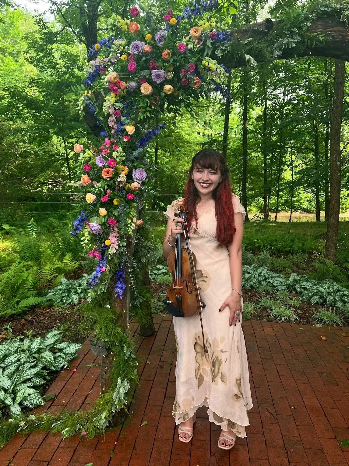 A woman with long red hair and a cream-colored dress with floral patterns holding a violin and bow, standing on a wooden pathway in a lush green outdoor setting, smiling in front of a large flower arrangement arch.
