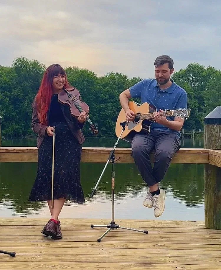A woman and a man playing musical instruments on a wooden dock by a lake. The woman is holding a violin and bow, smiling, wearing a brown leather jacket and a long dark skirt. The man is sitting on the edge of the dock, playing an acoustic guitar, wearing a blue shirt and dark pants. There is a microphone stand in front of them, and a background of trees and a cloudy sky.
