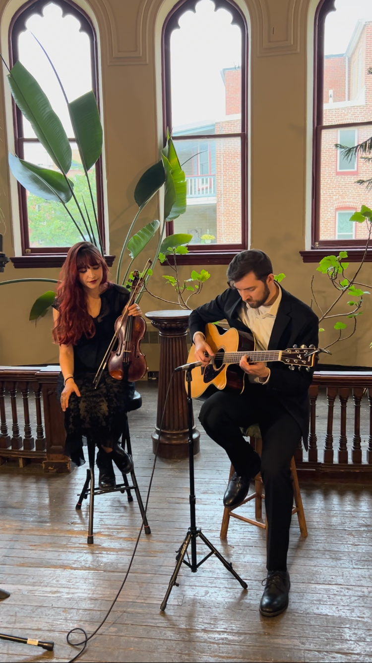 A man and woman performing music indoors with large arched windows and lush green plants behind them. The woman is holding a violin, and the man is playing an acoustic guitar.