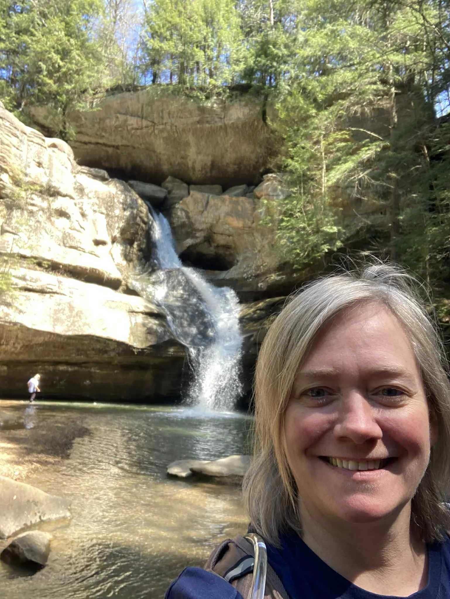 Woman with blonde hair smiling in front of a waterfall in a forested area.