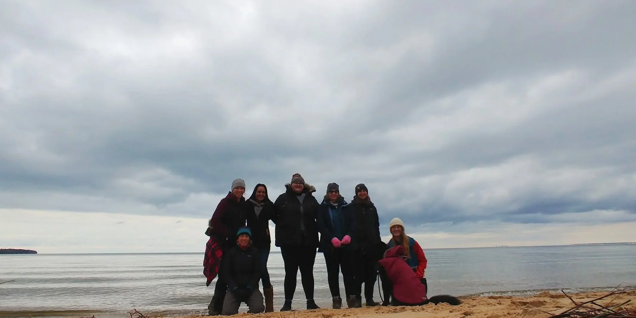Group of seven people standing on a sandy beach near the water, dressed warmly in jackets and hats, with cloudy sky overhead.