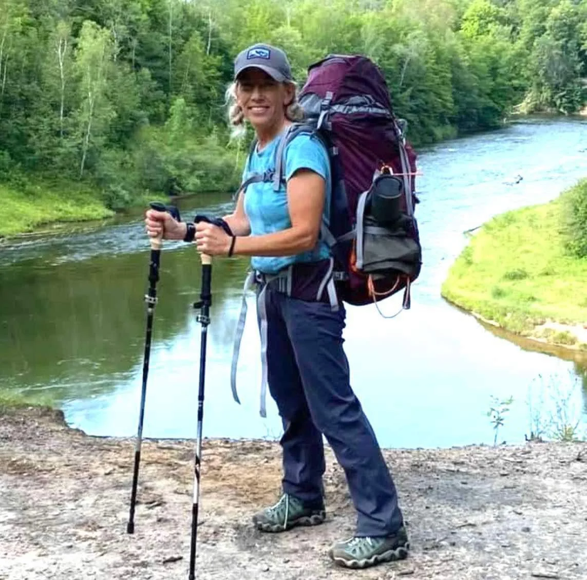 A woman standing on a dirt path near a river, wearing hiking gear, holding trekking poles, and smiling at the camera with a lush green forest in the background.