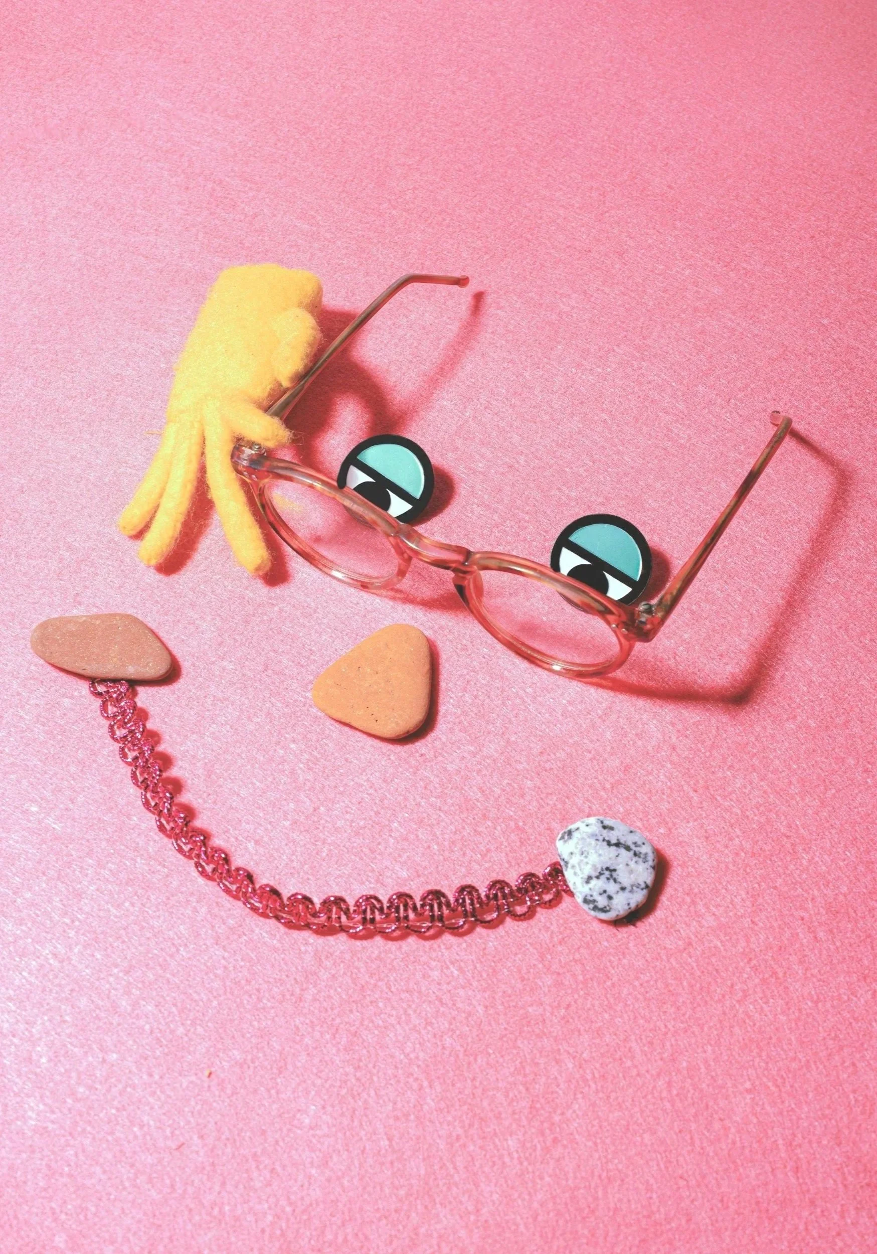 Pink background with a pair of pink sunglasses with cartoonish eye stickers, a yellow fuzzy octopus keychain, a pink chain necklace, and a small black and white speckled heart-shaped stone.