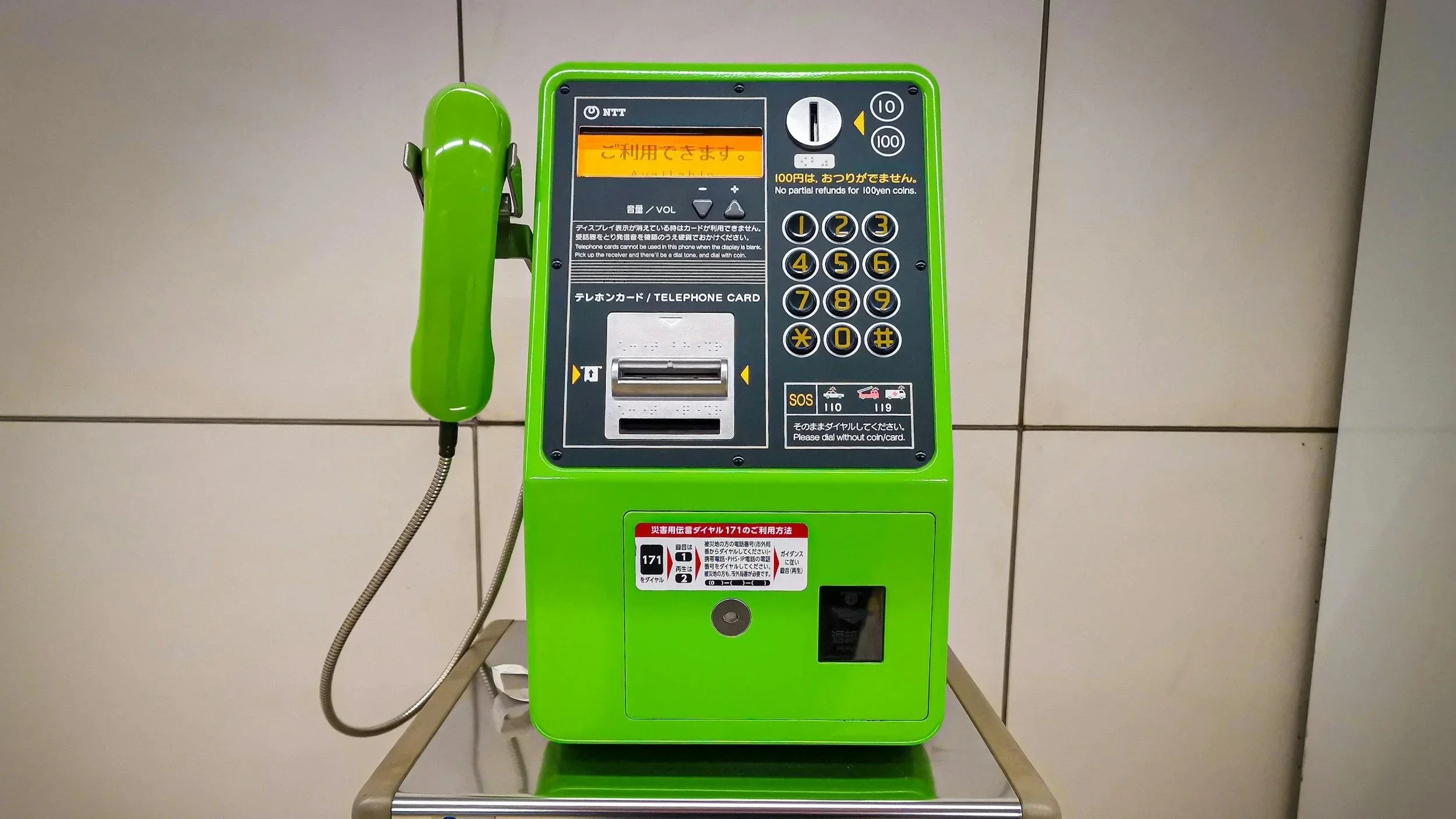 A bright green public telephone booth with a rotary dial and a green handset mounted on the side, set against a tiled wall.