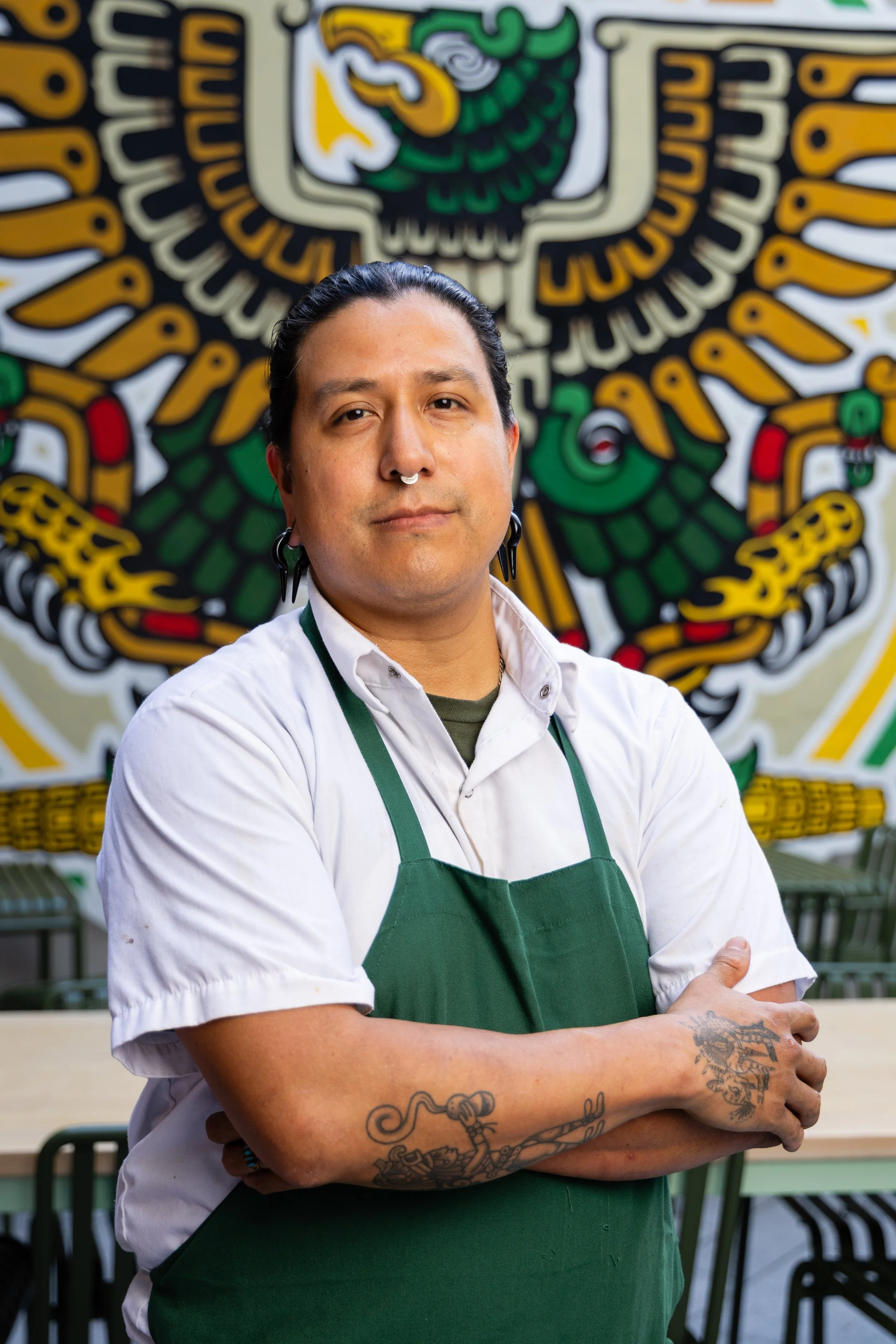 A person with dark hair, earrings, and a nose ring standing with arms crossed in front of a colorful mural of an eagle with Mexican motifs.