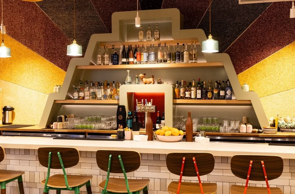 Bar with tiered shelves displaying liquor bottles, glasses, and decor, with six wooden chairs with green and red accents in front, and colorful textured wall panels.