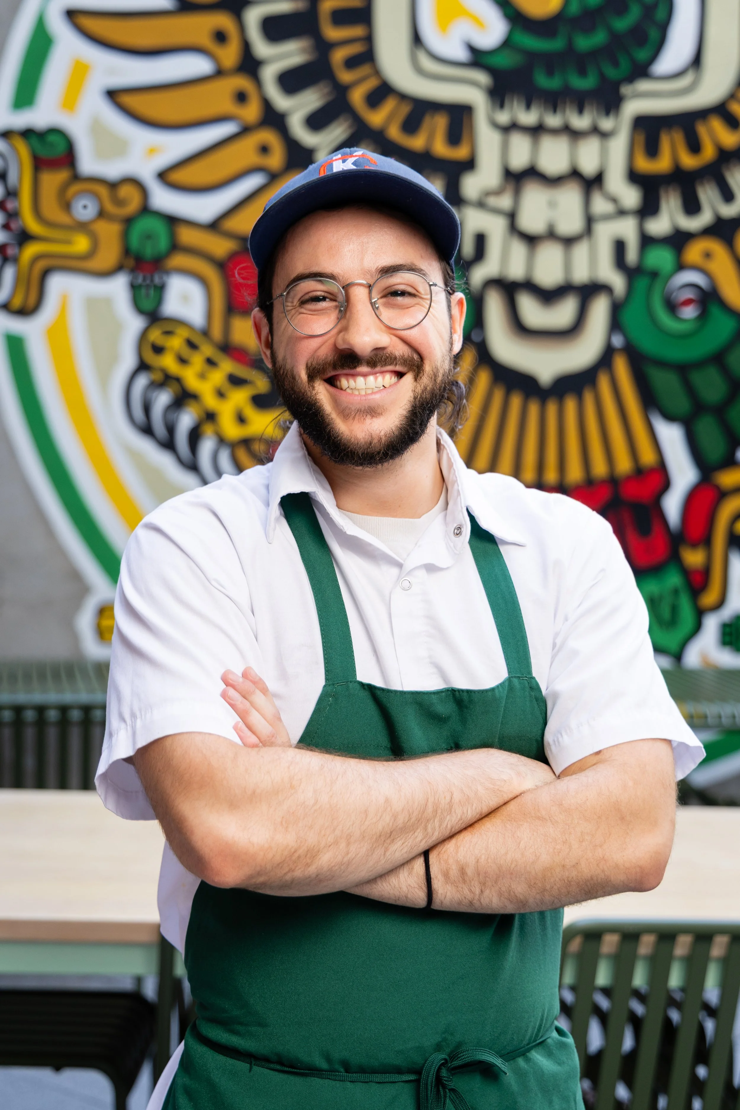 A smiling man with glasses, a beard, and a blue cap, standing with arms crossed in front of a colorful mural in a restaurant or café.
