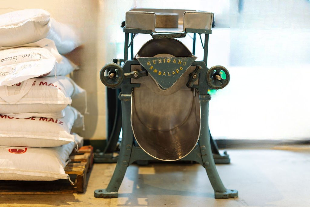 Vintage Mexican tortilla maker machine with bags of cornmeal on a wooden pallet to the left side.
