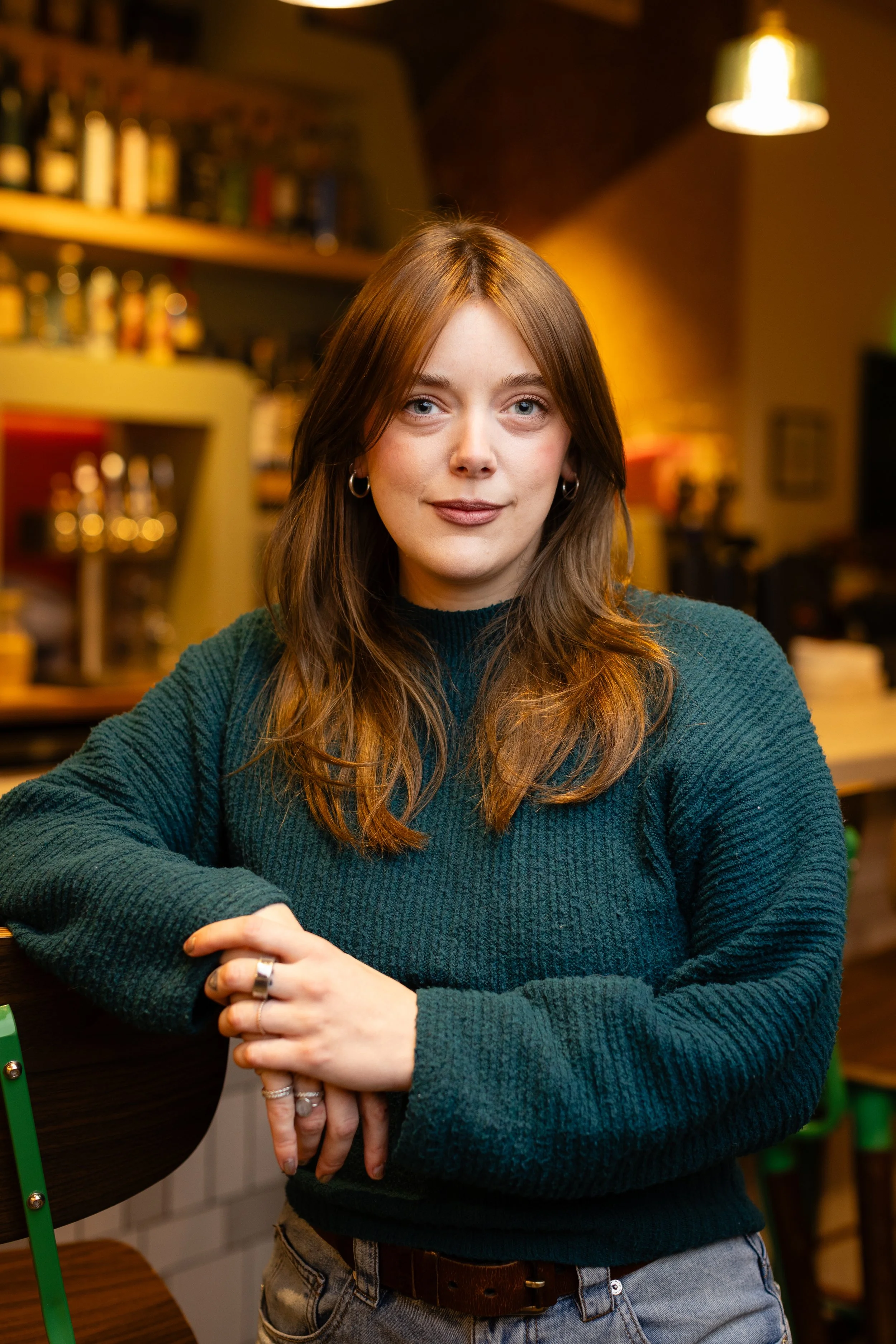 A young woman with long, wavy blonde hair, wearing a green button-up shirt and dark jeans, standing indoors near a window with a brick and concrete wall background.