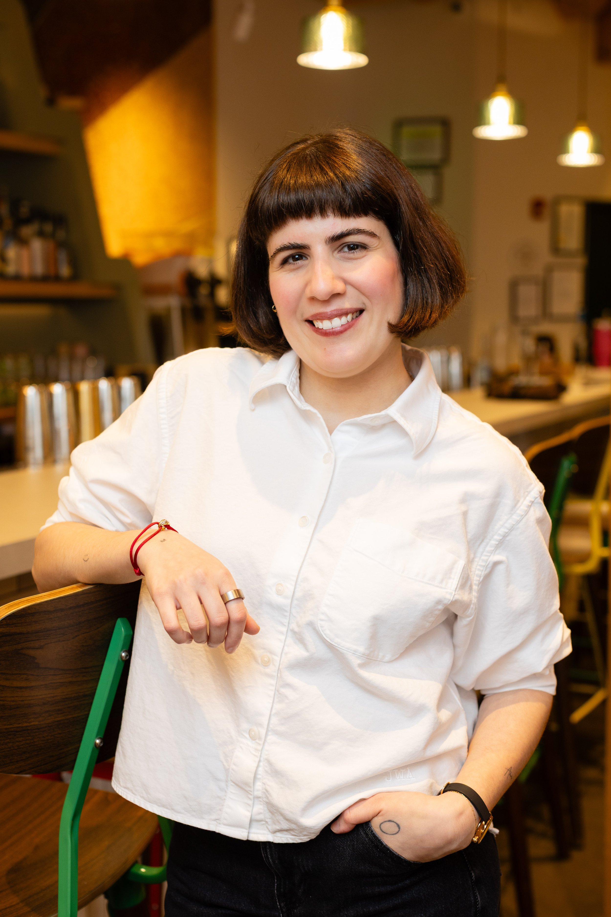 Woman with shoulder-length dark hair wearing round sunglasses and a colorful floral dress sitting on a pink outdoor chair outside a cafe with a pink window frame.