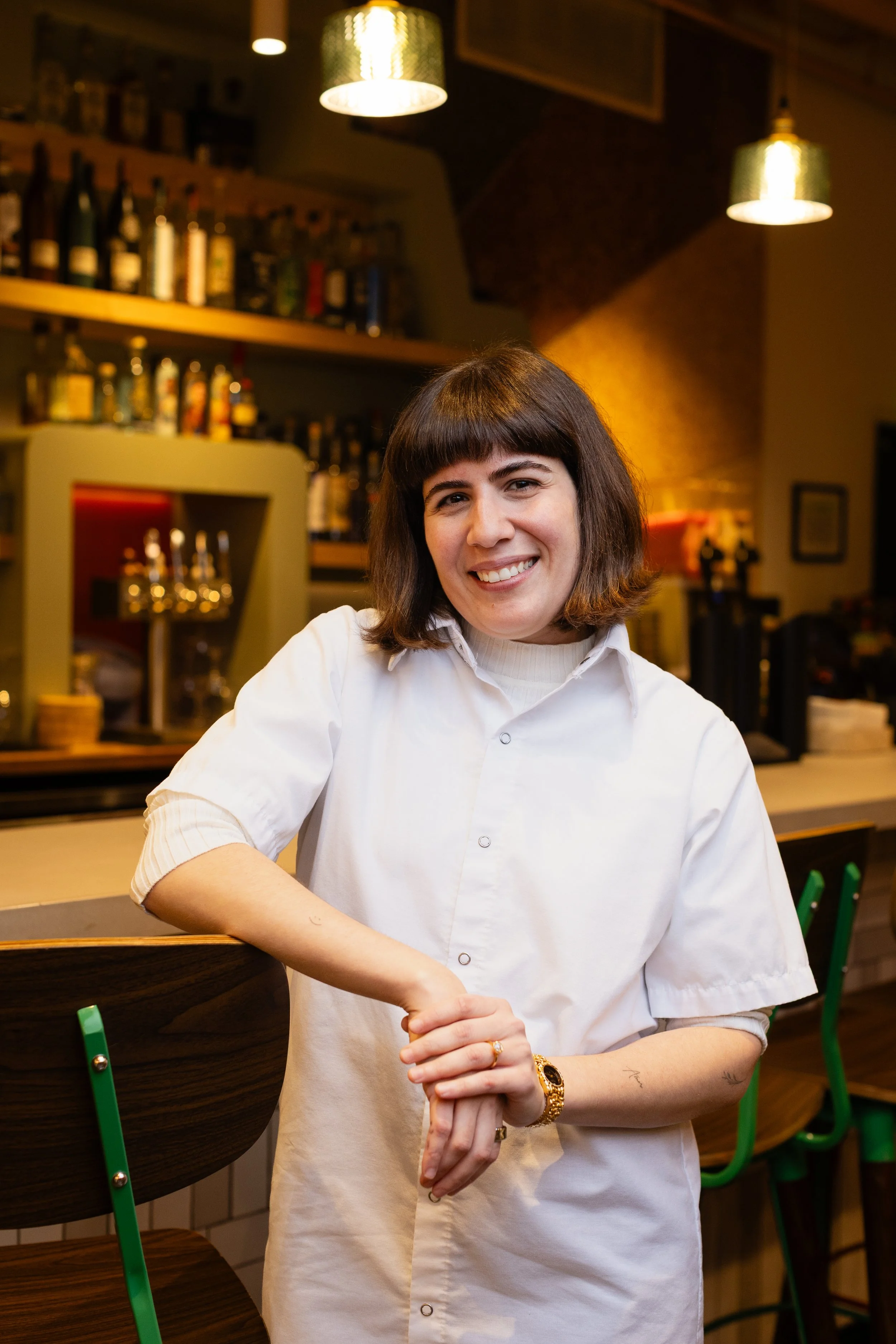 Woman with shoulder-length dark hair wearing round sunglasses and a colorful floral dress sitting on a pink outdoor chair outside a cafe with a pink window frame.