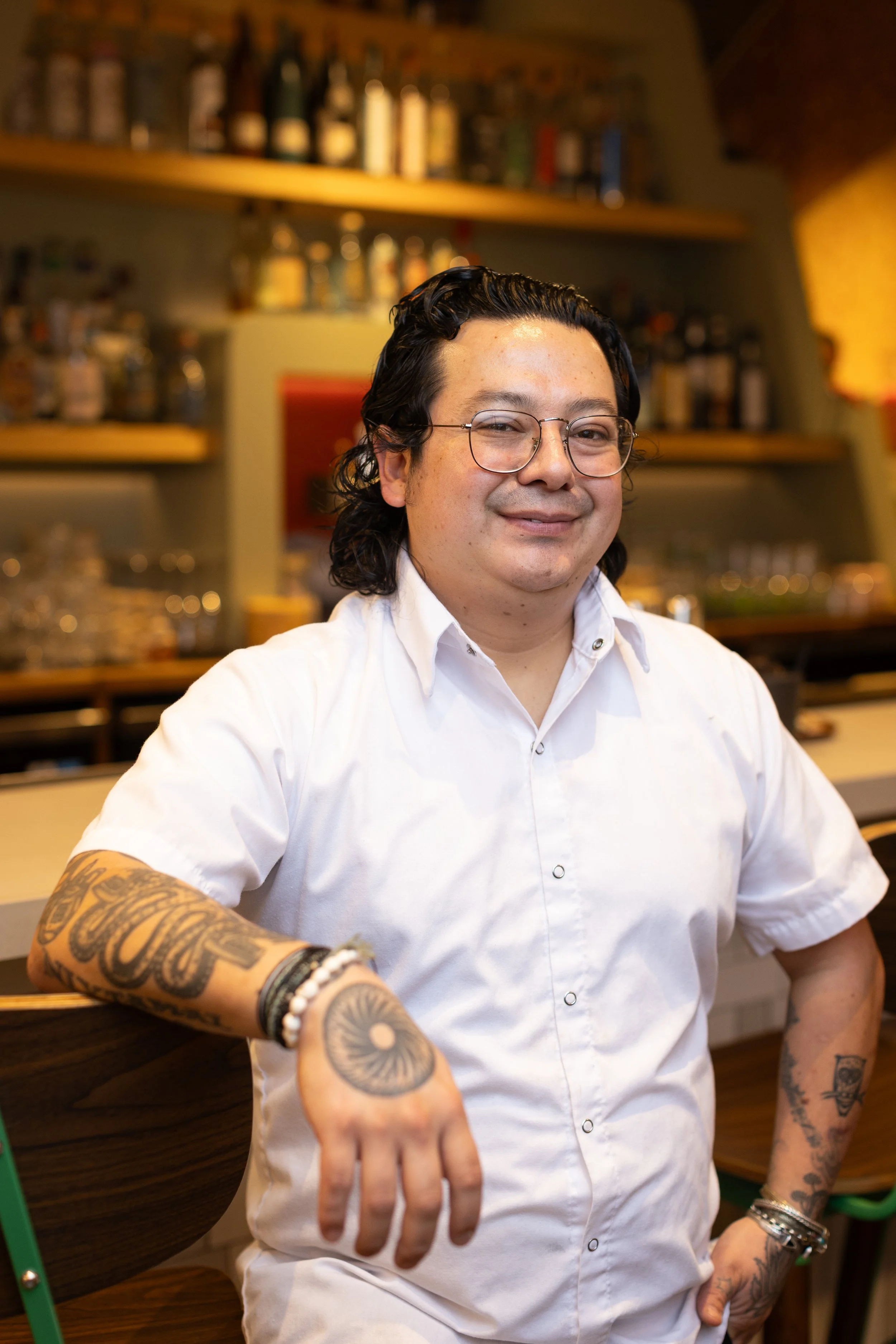 A smiling man with glasses, tattoos on his arms, and wearing a green apron over a white shirt, standing in front of a colorful mural.