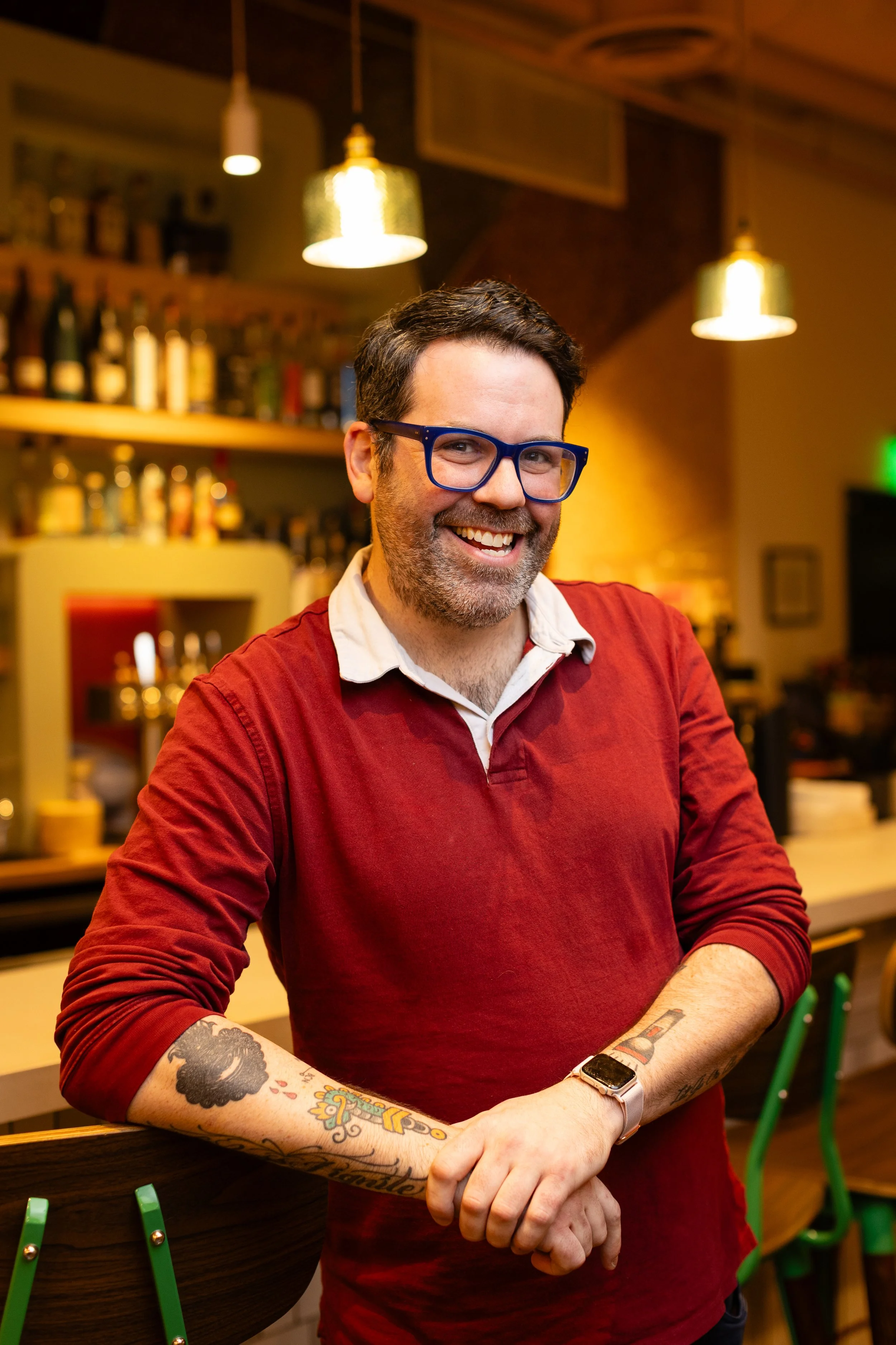 A smiling man with glasses and a beard standing with arms crossed inside a restaurant. The background shows a bar area with stools and a sign that says 'MAIZ'.