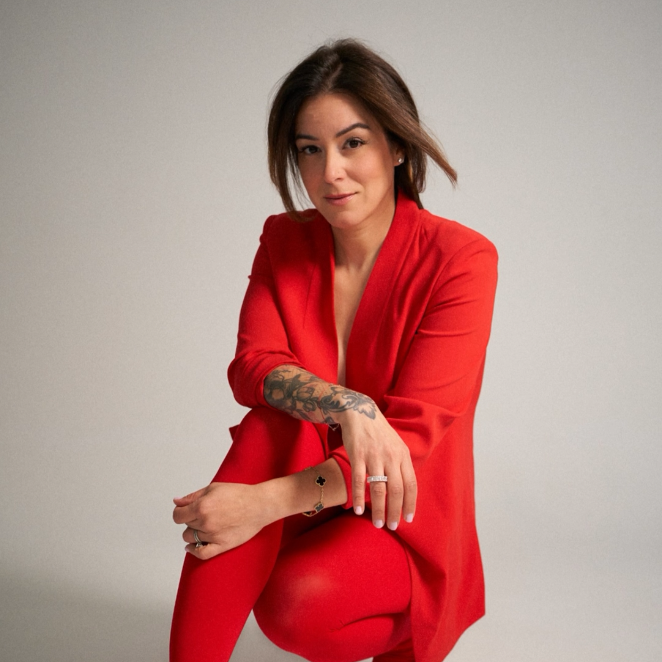A woman with short brown hair wearing a red outfit, sitting with her legs crossed, posing in front of a plain grey background.