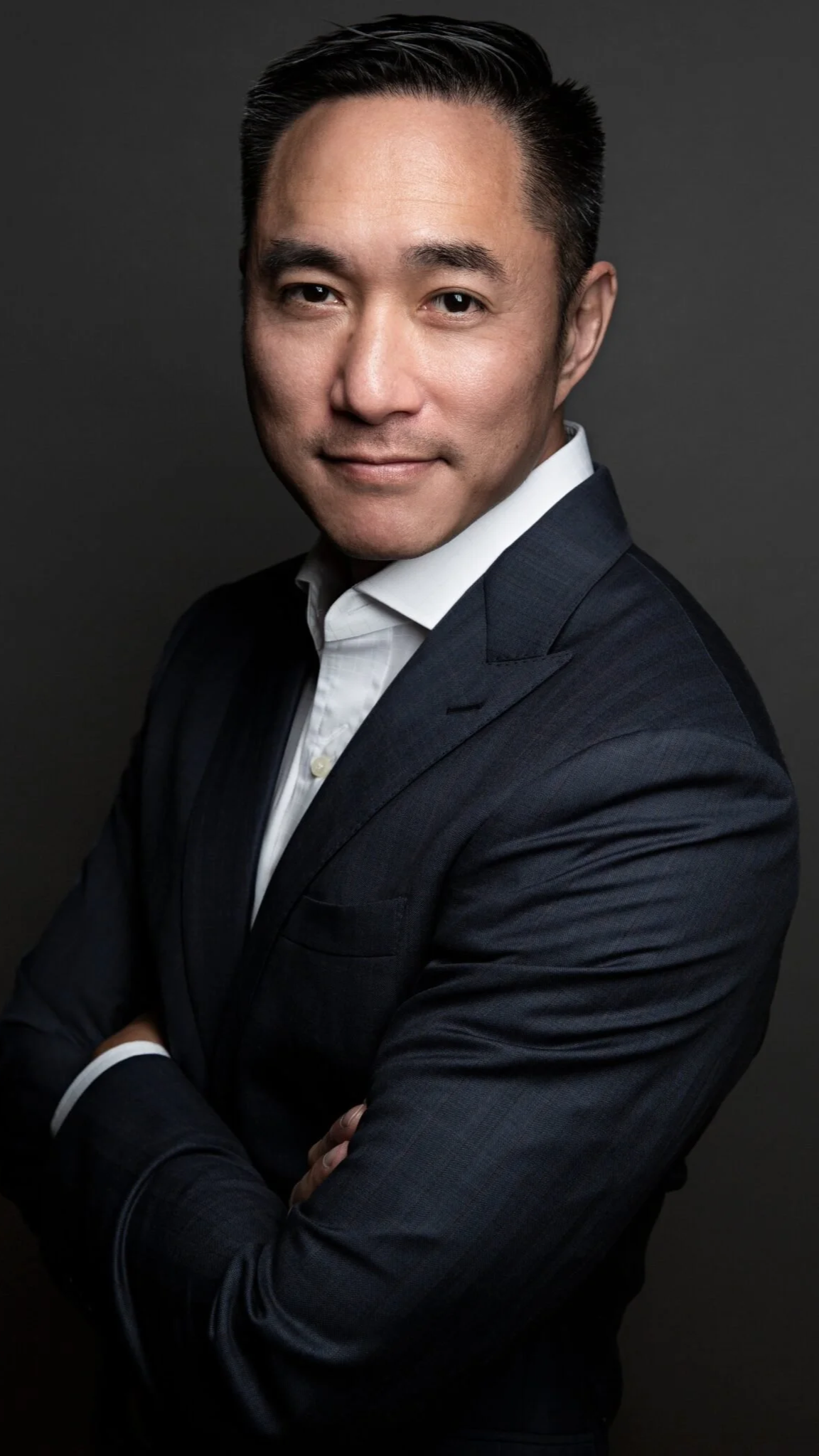 Professional portrait of a man in business attire with crossed arms, dark suit, white shirt, against gray background.