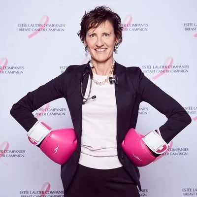 A woman with short dark hair wearing a black blazer, white top, and a pearl necklace, standing with hands on hips in pink boxing gloves at a breast cancer awareness event.