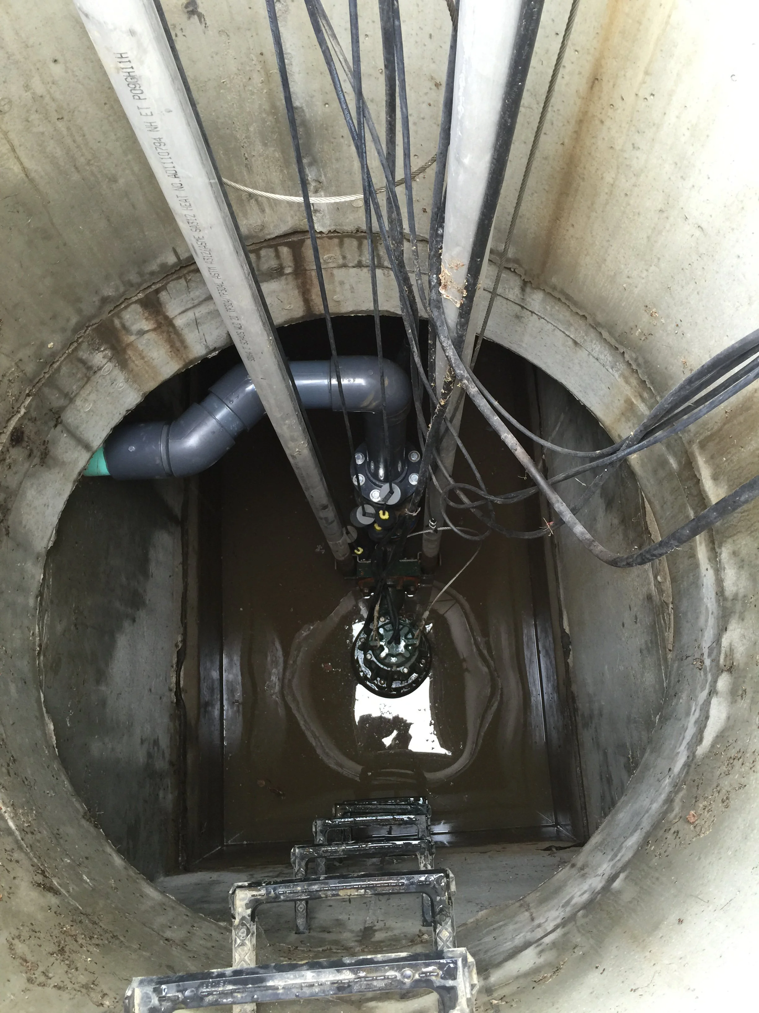 View inside a large underground shaft showing pipes, electrical cables, and a water-filled bottom with a ladder leading down.