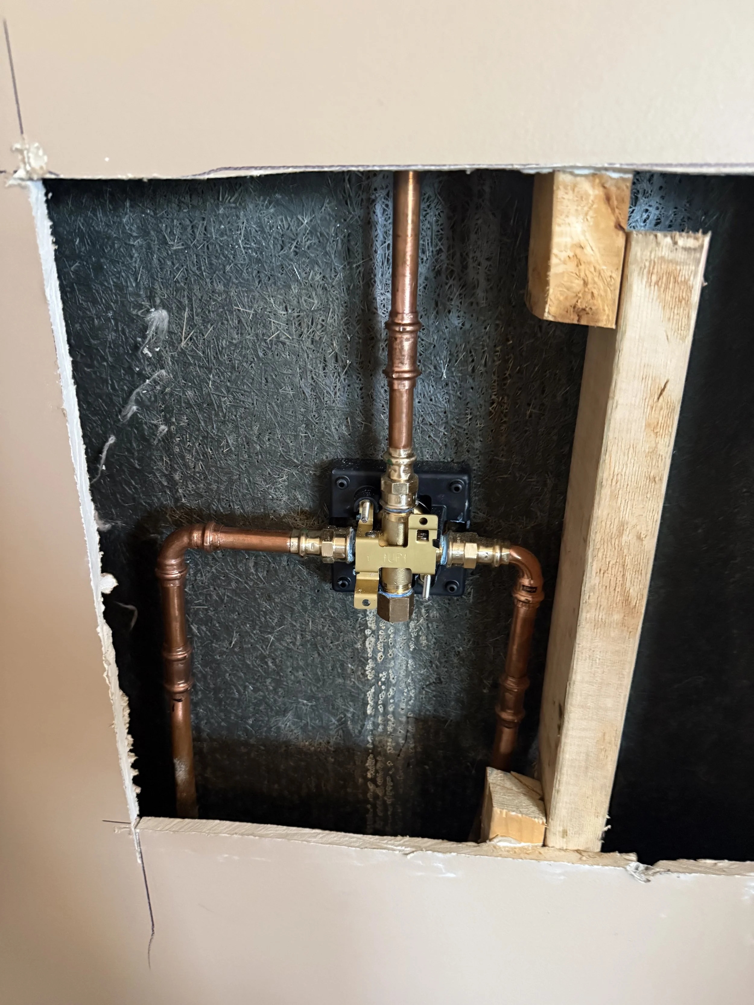 View of an unfinished wall with plumbing pipes and a valve through a square opening in the drywall, with wooden framing visible.