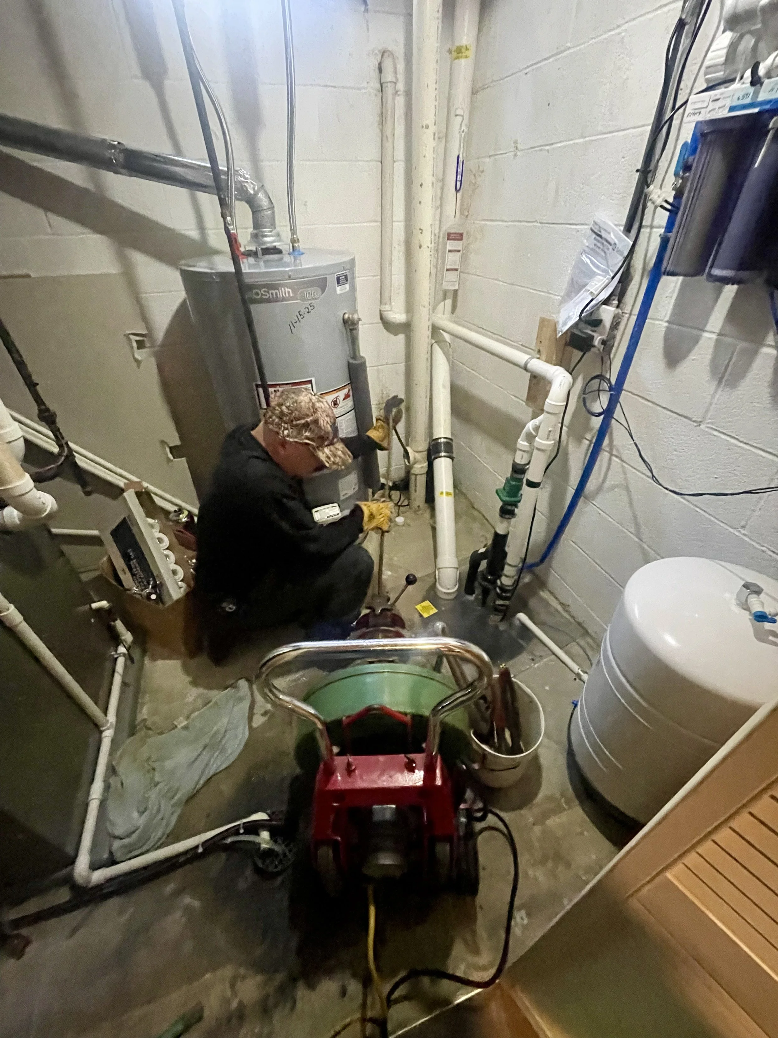 A plumber working on pipe connections near a water heater in a basement utility room, with a sump pump and various tools around him.